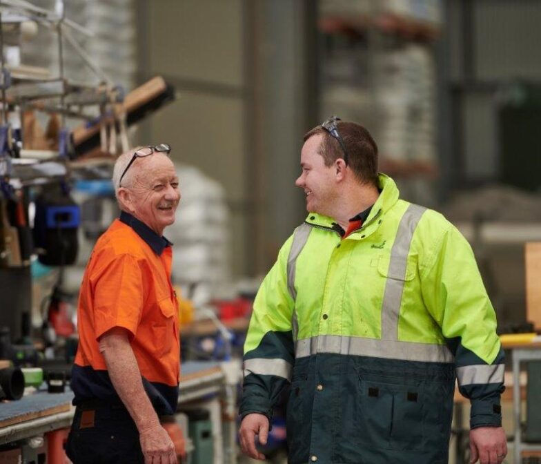 Two Polymaster factory workers in hi-vis clothing smiling and talking together on the warehouse floor surrounded by manufacturing equipment and fittings
