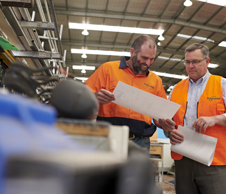 Two Polymaster team members in branded orange hi-vis vests reviewing production documents together on the factory floor