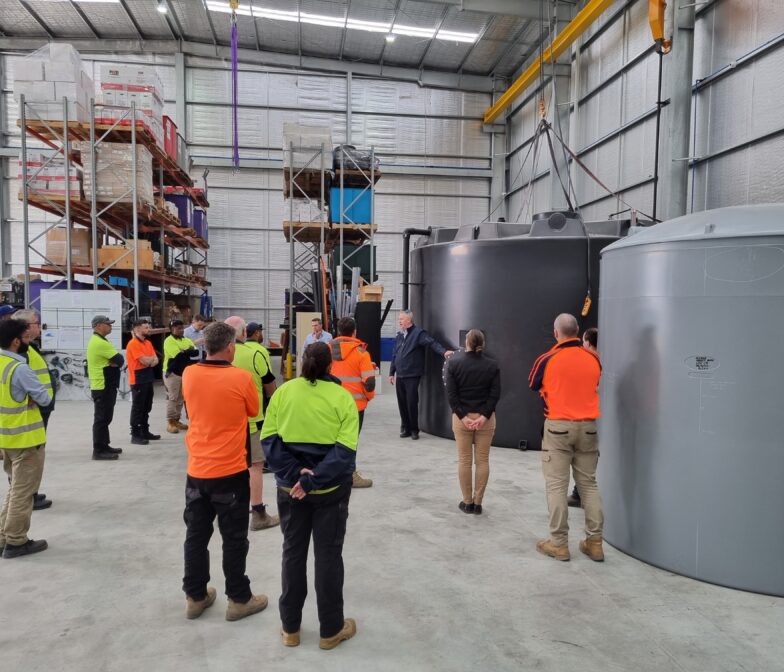 Polymaster team members standing in a warehouse beside large water tanks during a factory briefing